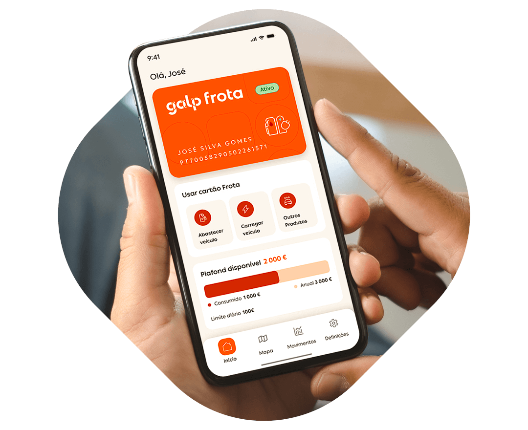 Fotografia de duas mãos a segurar num telemóvel. No ecrã é visível a página inicial da app Galp Frota, com o cartão no topo, seguido de um menu, seguido do plafind disponível, seguido do menu da aplicação.