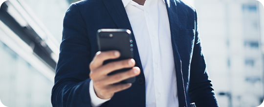Fotografia em grande plano de uma mão a mexer no telemóvel. Nota-se que a pessoa veste uma camisa branca e um blaser azul-escuro.