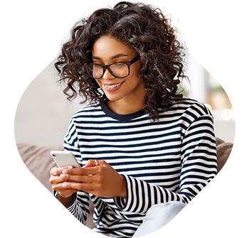 Fotografia de uma senhora. Está a mexer no telemóvel com as duas mãos. Veste uma camisola às riscas pretas e brancas de manga comprida. Tem o cabelo escuro, encaracolado, e usa óculos de vista.
