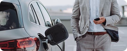 Fotografia de um carro cinzento a ser carregado, com um homem ao seu lado direito, vê-se a cintura e o peito. Veste um fato: camisa branca, e calça e blaser cinza claro, quadriculado.