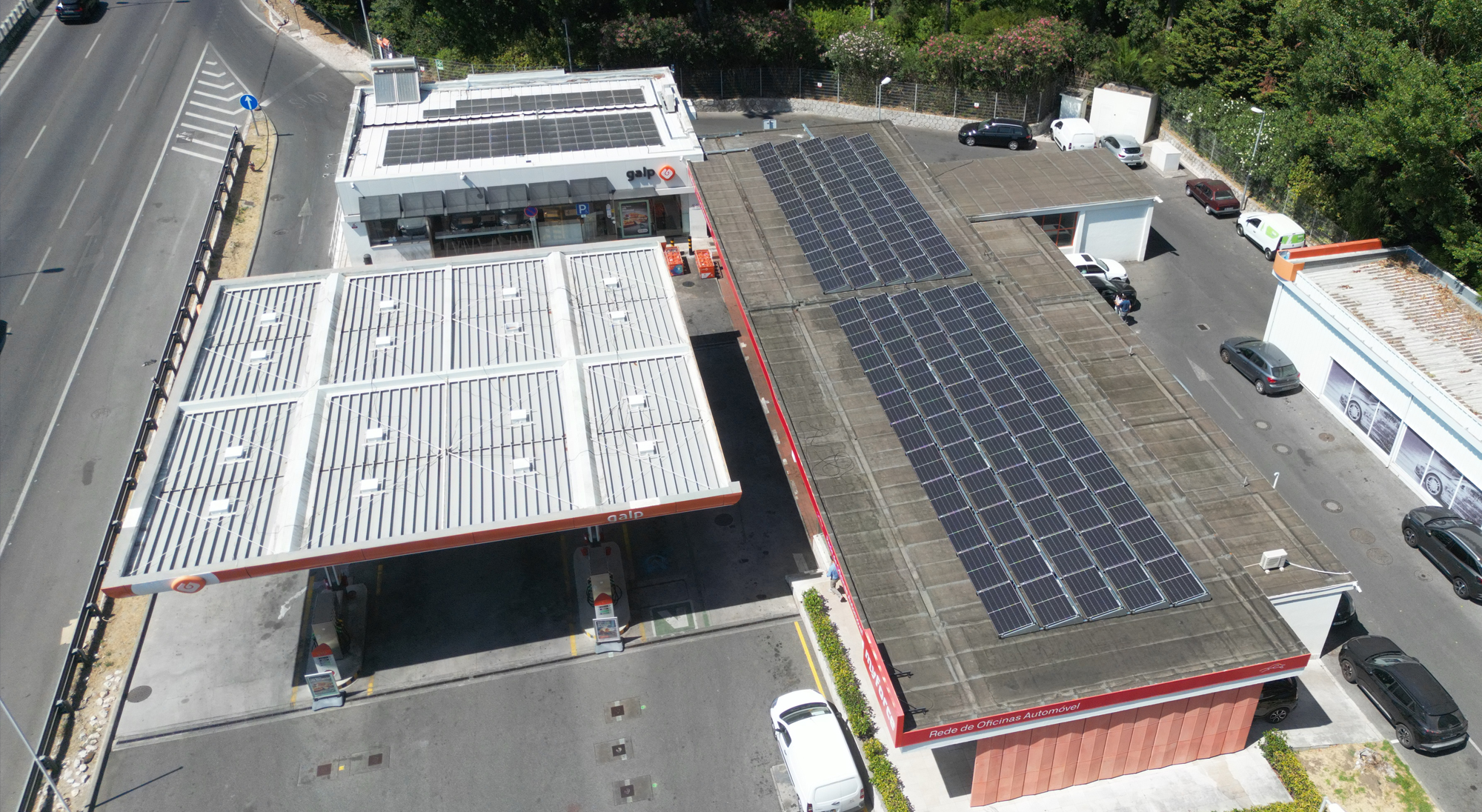 Vista aérea de um posto de combustível Galp com painéis solares nos telhados, carros estacionados e a circular, e áreas verdes circundantes.