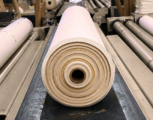 Rolo de tecido em processo de fabrico numa unidade industrial. A imagem representa produção têxtil, transformação de materiais e operações industriais de acabamento e processamento.