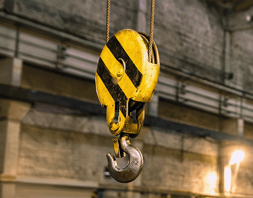 Gancho de elevação industrial suspenso numa instalação fabril. A imagem representa equipamentos de movimentação de cargas, engrenagens abertas e manutenção industrial.