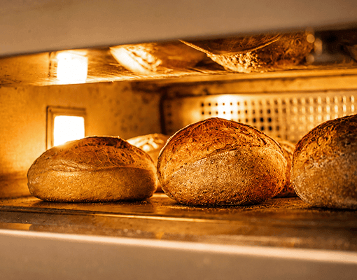 Pães em processo de cozedura num forno industrial. A imagem representa produção alimentar, processamento industrial de alimentos e operações de panificação.