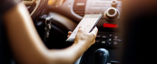 Fotografía en primer plano de una mano sosteniendo un teléfono celular. Dentro de un auto.