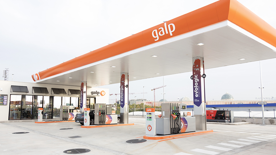 Posto de combustível Galp moderno, com cobertura laranja e logótipo visível no topo. Várias bombas de abastecimento estão organizadas sob a estrutura, incluindo zonas para veículos elétricos. Um carro está estacionado junto a uma das bombas. O espaço é amplo, limpo e iluminado, com edifícios urbanos e uma estrada ao fundo.