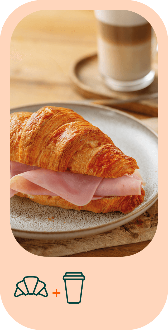 Fotografia em grande plano de um croissant com fiambre num prato. Atrás é visível uma caneca de galão.