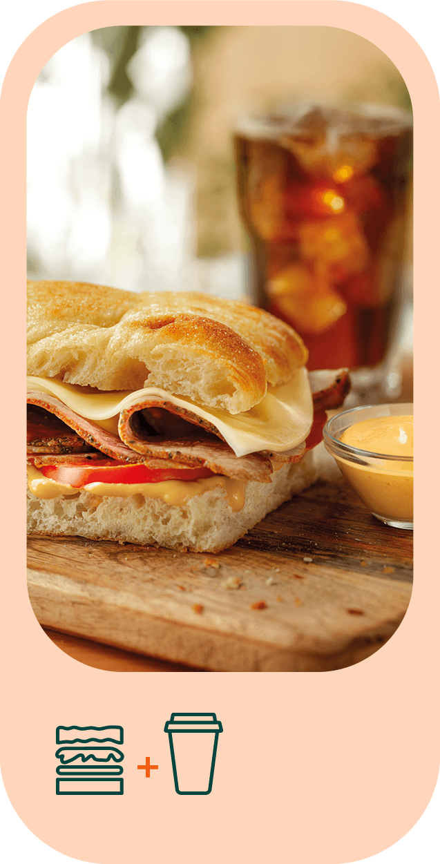 Fotografia em grande plano de uma focaccia cortada a meio. No seu interior é visível pastrami, queijo, tomate e molho. Atrás da mesma, vê-se um copo de sumo.